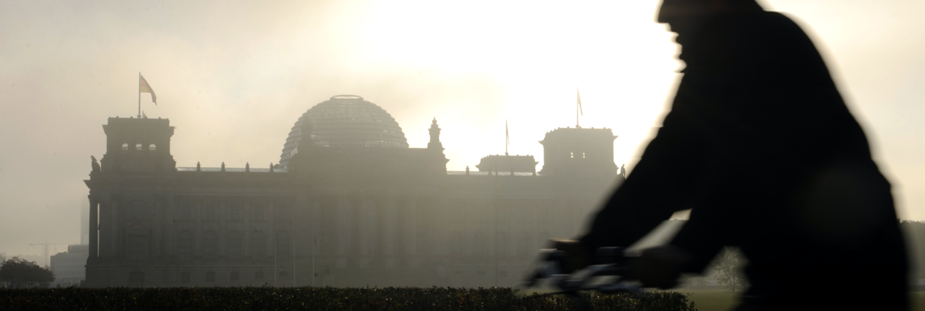 Silhouette eines Fahrradfahrers vor dem Reichstagsgebäude in Morgendunst mit dahinter aufgehender Sonne © picture-alliance/dpa/Rainer Jensen