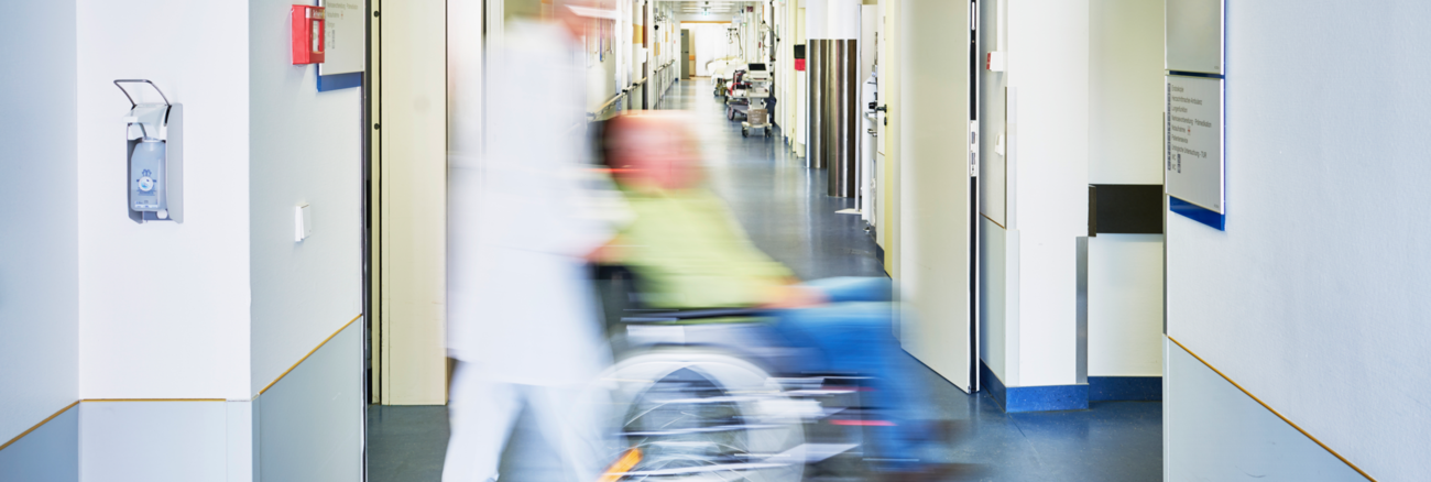 Ein Pfleger schiebt in einem Krankenhaus einen Patienten im Rollstuhl durch einen Flur mit einem Schild Notaufnahme an der Wand © iStock/upixa