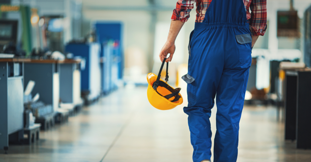 Ein Arbeit in blauer Latzhose und mit gelbem Helm in der Hand läuft durch eine Werkhalle © iStock/gilaxia