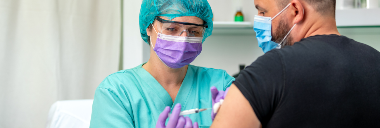 Krankenschwester mit Schutzbrille und Maske verabreicht einem Mann mit Maske eine Injektion @ iStock/zoranm