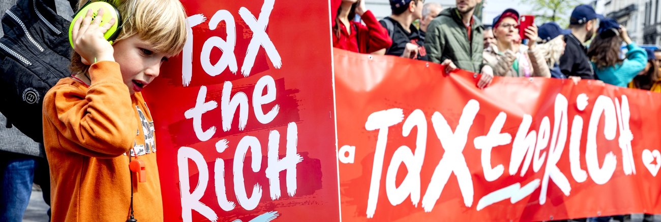 Teilnehmer an einer Demonstration halten ein Transparent und ein Plakat mit der Aufschrift: Tax the rich © picture alliance/dpa/Belga|Hatim Kaghat