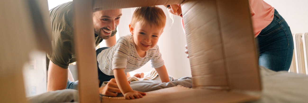 Ein Junge krabbelt durch einen Umzugskarton, den seine Eltern halten © iStock/AleksandarNakic