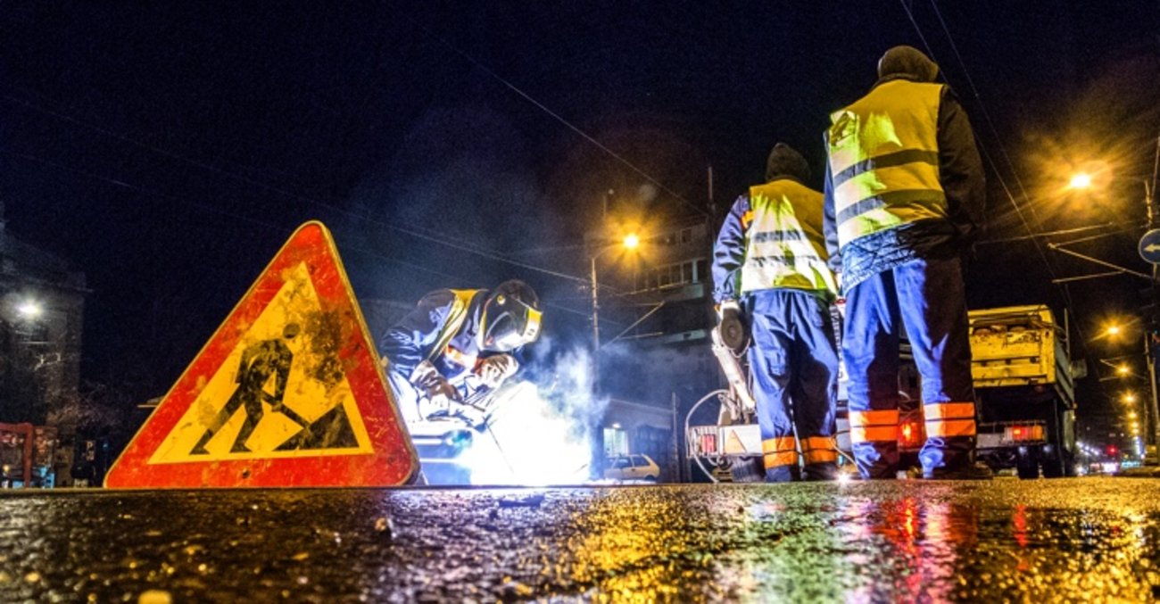 Ein Schweißer und zwei Arbeiter hinter einem Baustellen-Schild im Dunkeln bei Straßenbauarbeiten © iStock/EXTREME-PHOTOGRAPHER