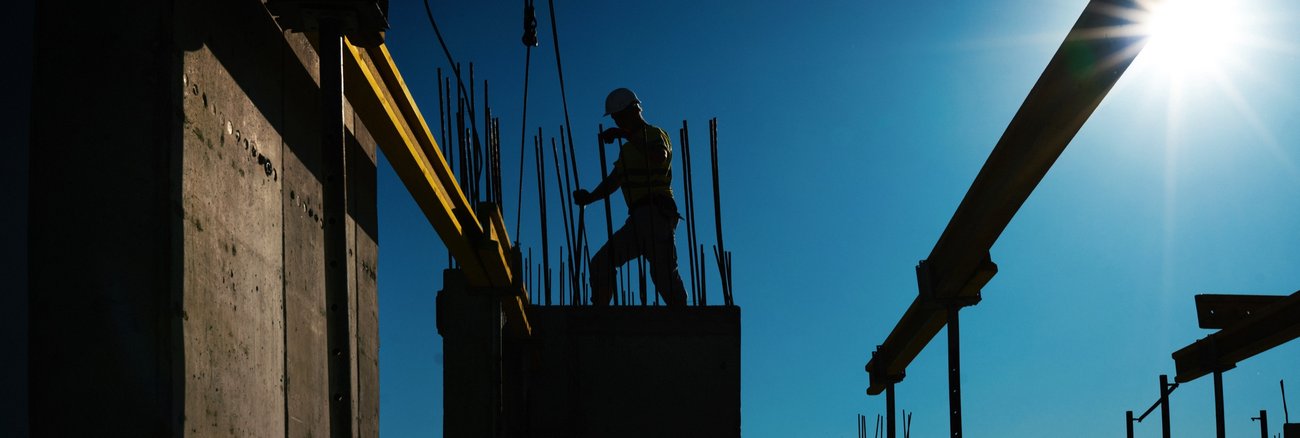 Silhouette eines Bauarbeiters auf einer Baustelle mit Stahlträgern, Kran und Betonpfeilern © iStock/gilaxia