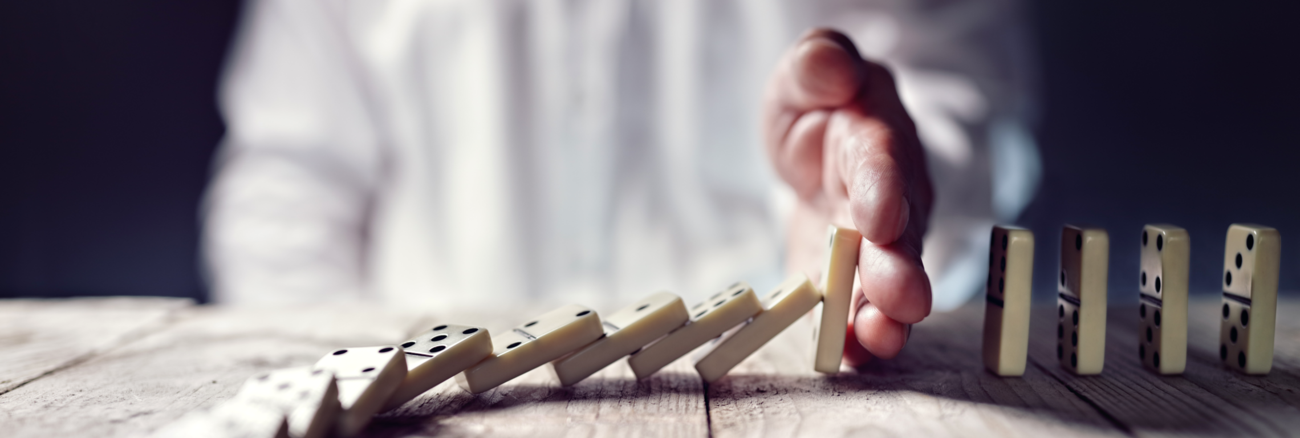 Ein Mann stoppt mit seiner Hand das Umfallen einer Domino-Kette © iStockphoto.com/BrianAJackson