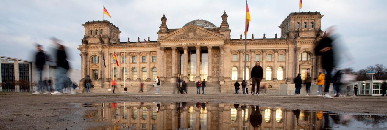 Das Pleargebäude des Bundestages spiegelt sich in einer Pfütze © picture alliance/dpa/Christoph Soeder