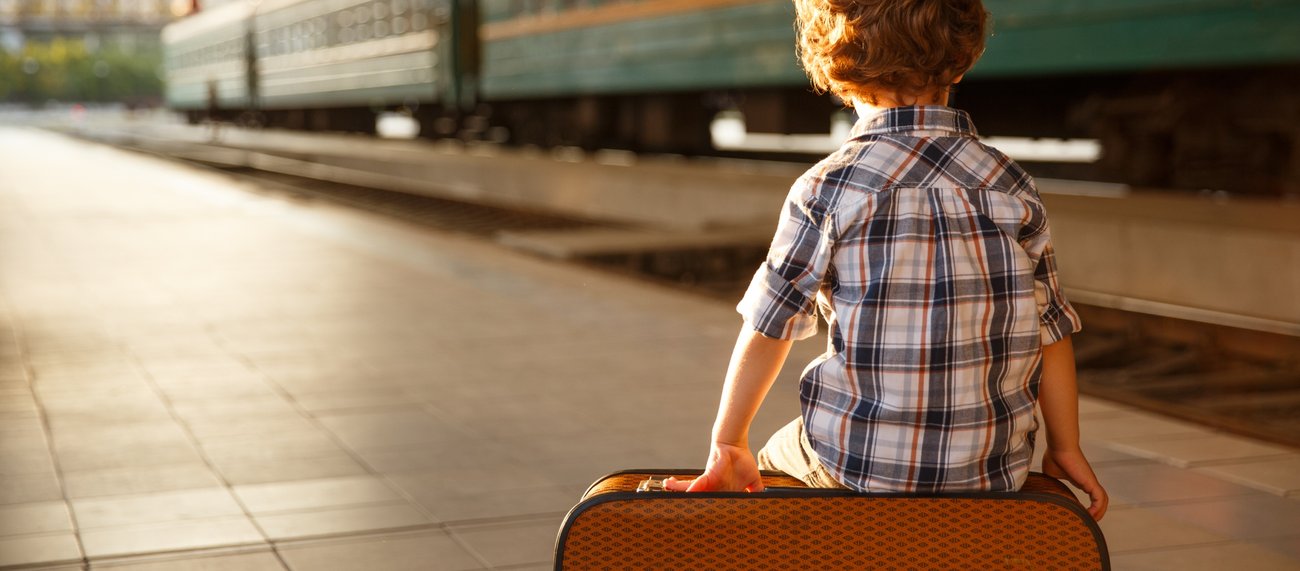 Ein Junge allein auf einem Bahnsteig vor einem Zug sitzt auf einem Koffer @ iStock/max-kegfire