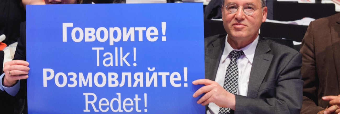 Gregor Gysi hält ein blaues Schild mit der Aufschrift »Redet!« in Russisch, Englisch, Ukrainisch und Deutsch