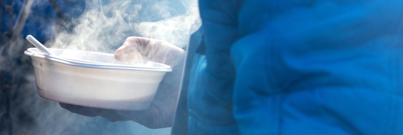 Eine Person mit blauer Jacke hält eine Plastikschale mit dampfender Suppe © iStock/wjarek