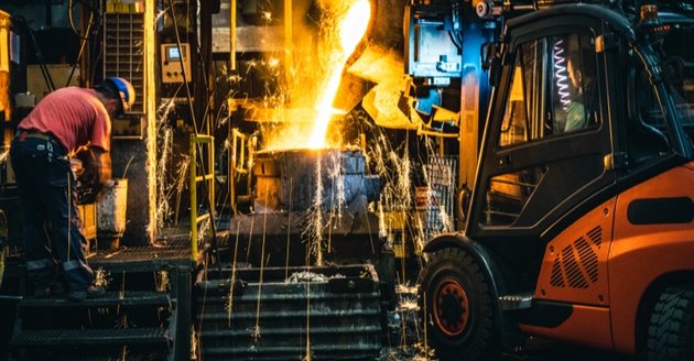 In einem Stahlwerk wird geschmolzenes Metall in eine Gießform gegossen, daneben eine Arbeiter und ein Gabelstapler © iStock/simonkr