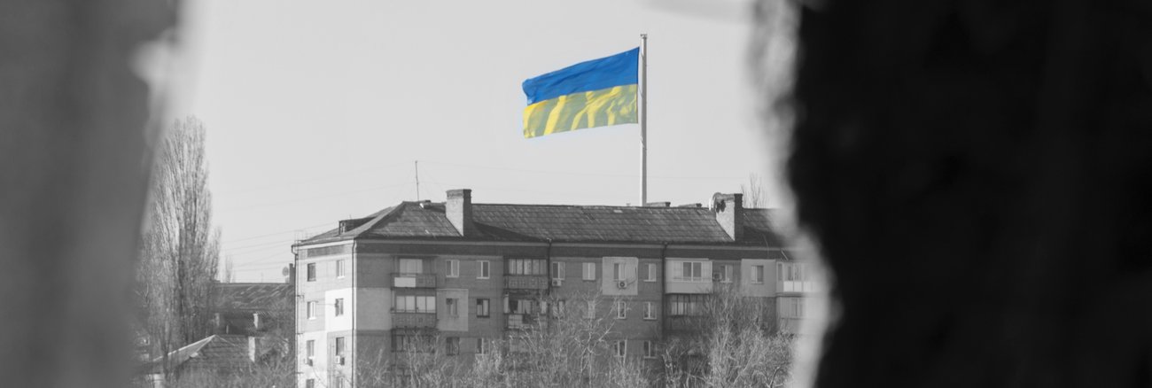 Blick durch ein Fenster eines zerstörten Hauses auf die ukrainische Flagge auf einem gegenüberliegenden Haus © iStock/Ihor Martsenyuk