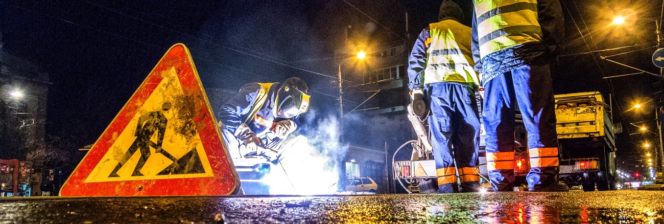 Ein Schweißer und zwei Arbeiter hinter einem Baustellen-Schild im Dunkeln bei Straßenbauarbeiten © iStock/EXTREME-PHOTOGRAPHER