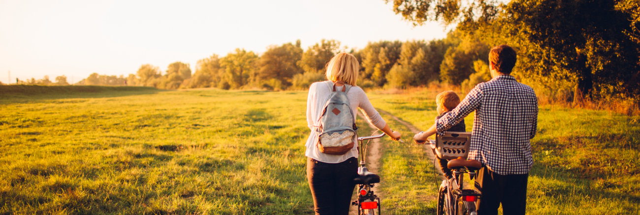 Junge Eltern mit Kind schieben zwei Fahrräder über ein Feld © iStock/AleksandarNakic Junge Eltern mit Kind schieben zwei Fahrräder über ein Feld © iStock/AleksandarNakic