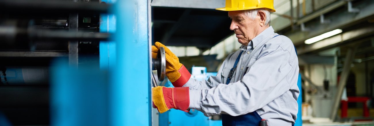 Ein älterer Arbeiter mit gelbem Schutzhelm schraubt an einer blauen Maschinen © iStock/SeventyFour