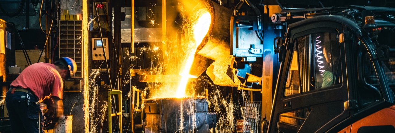 In einem Stahlwerk wird geschmolzenes Metall in eine Gießform gegossen, daneben eine Arbeiter und ein Gabelstapler © iStock/simonkr
