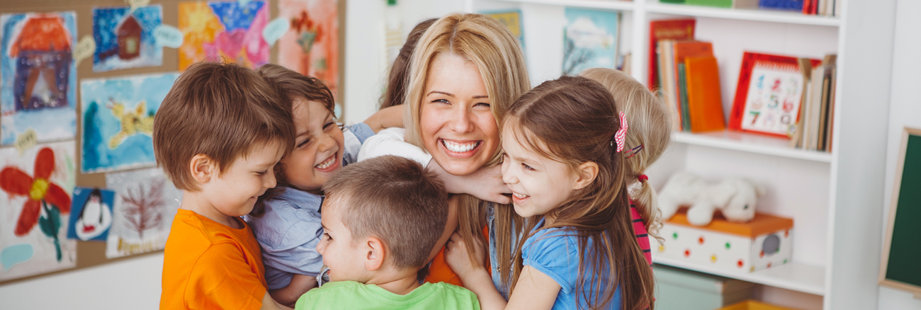 eine junge Erzieherin wird von einer Gruppe Kita-Kinder umarmt und lacht. Foto: © istock.com/svetikd