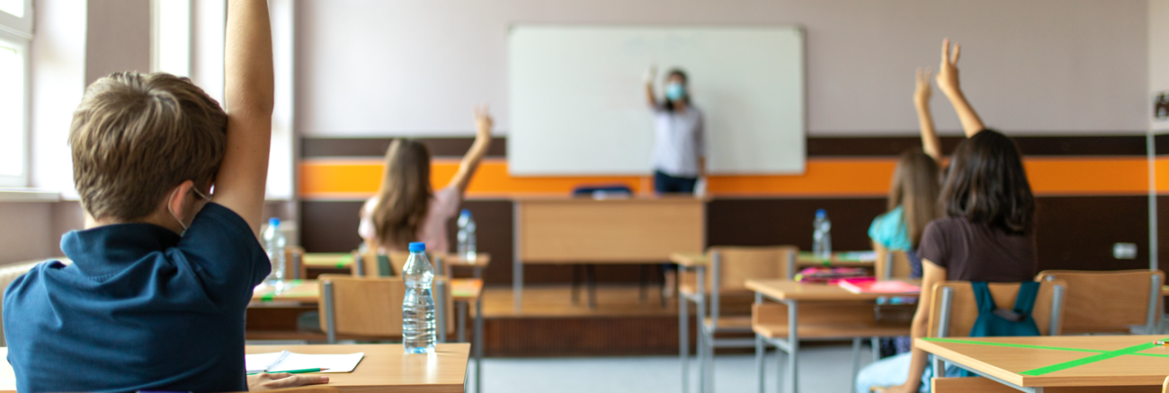 Schüler:innen mit Masken in einem Klassenzimmer melden sich, im Hintergrund eine Lehrerin mit Maske vor der Tafel © iStock/miljko