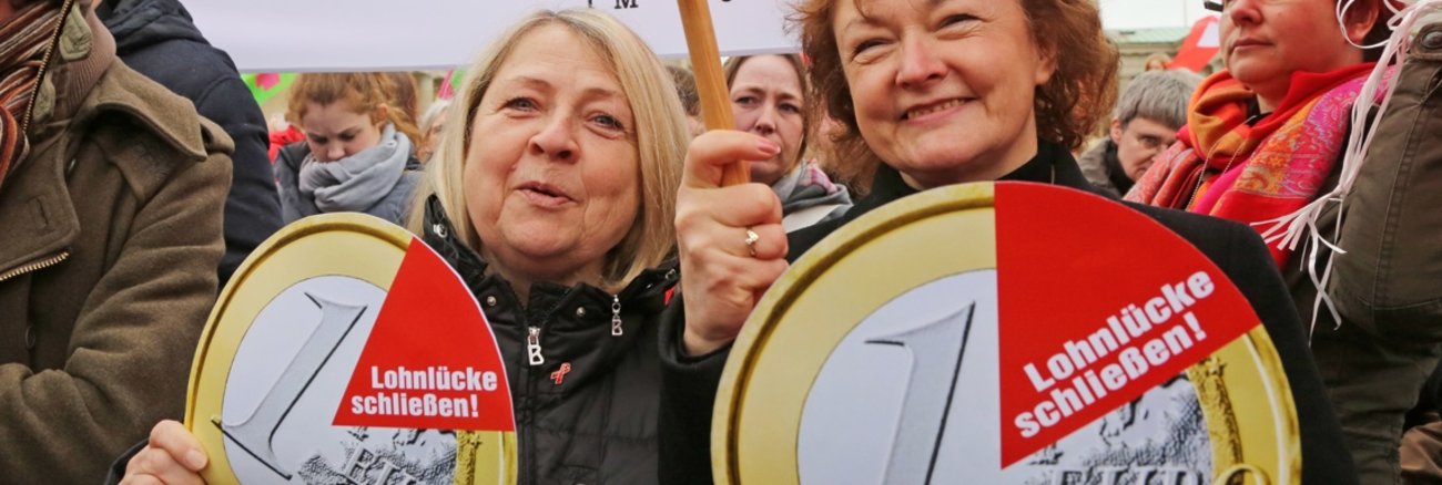 Birgit Wöllert und Kerstin Kassner halten am Equal Pay Day Schilder in Form einer Ein-Euro-Münze mit der Aufschrift: Lohnlücke schließen