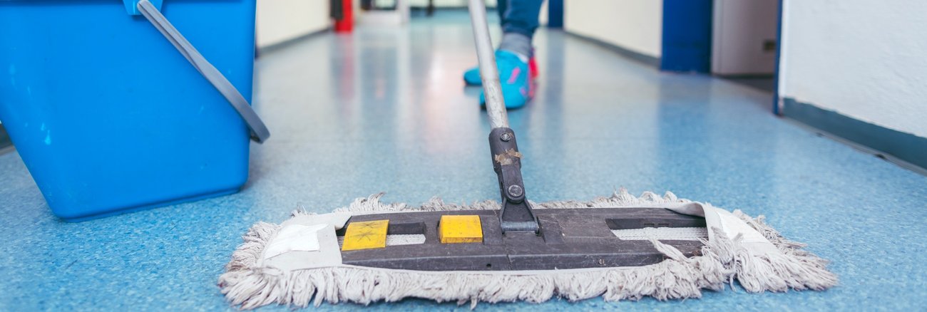 Eine Reinigungskraft wischt mit einem Wischmob den Fußboden in einem Büroflur © iStock/kzenon