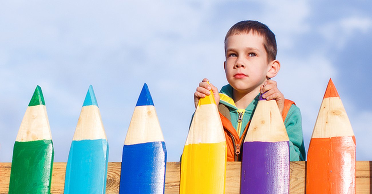 Kind blickt nachdenklich über einen Zaun, dessen Latten wie Buntstifte aussehen. Foto: © istock.com/EvgeniiAnd