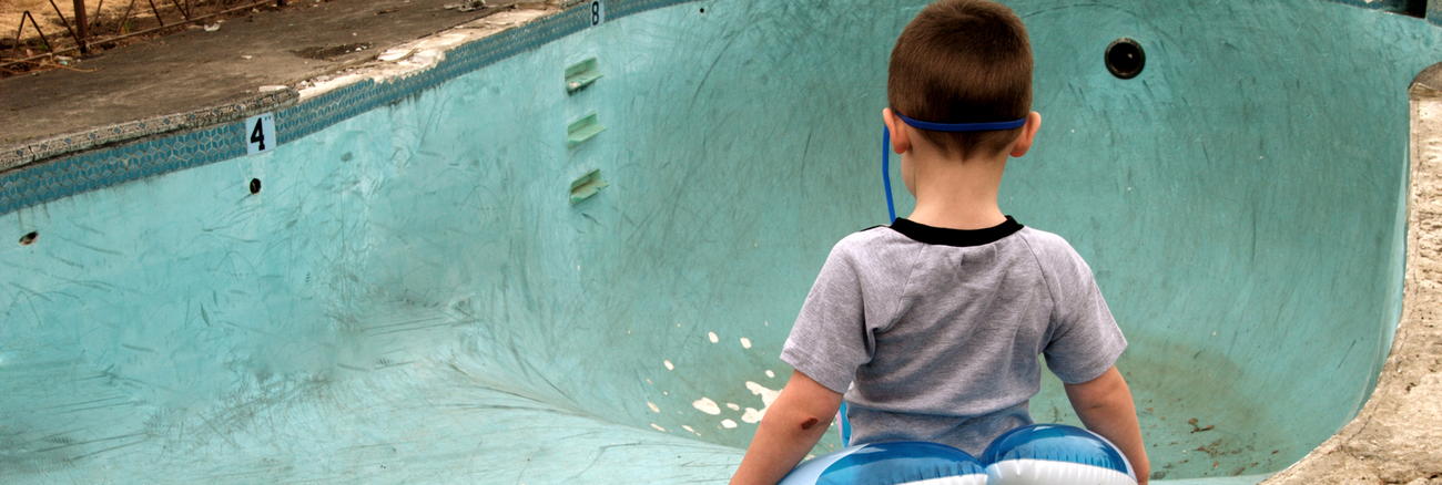 Ein kleiner Junge mit Schwimmhilfe steht vor einem leeren, renovierungsbedürftigen Schwimmbecken in einem Freibad. Foto: © iStock.com/RickLordPhotography