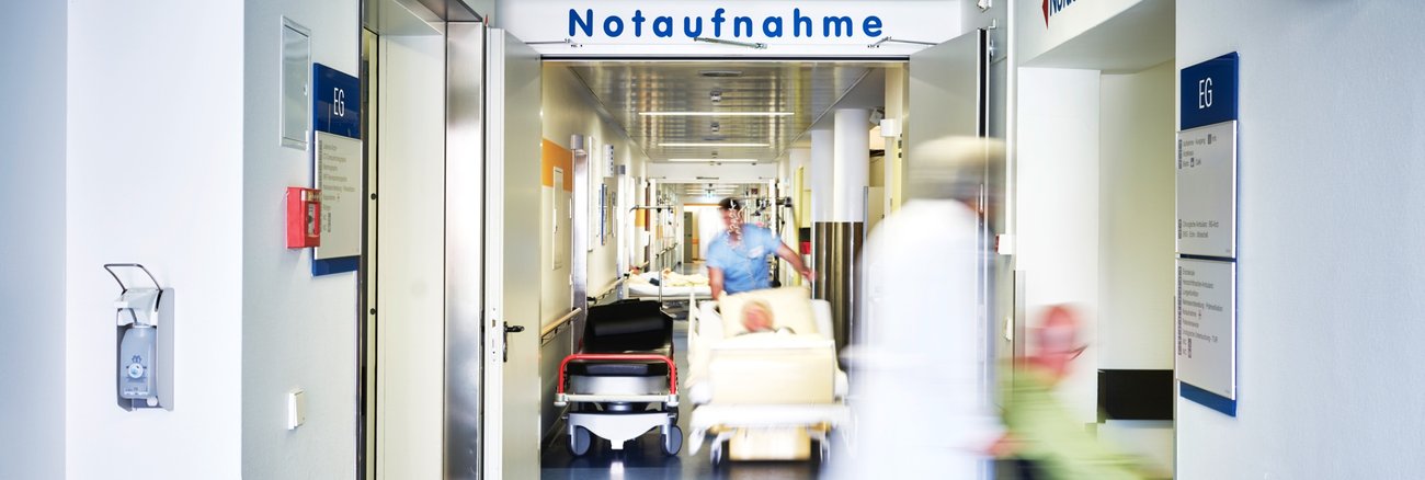 Krankenpfleger und Arzt schieben Rollstuhl und Fahrtrage in einem Krankenhausflur, darüber ein Schild mit Aufschrift Notaufnahme © iStocj/upixa