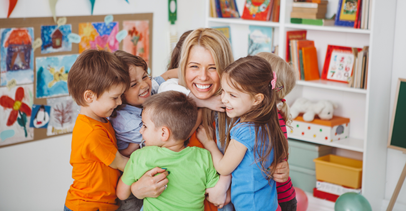 eine junge Erzieherin wird von einer Gruppe Kita-Kinder umarmt und lacht. Foto: © istock.com/svetikd