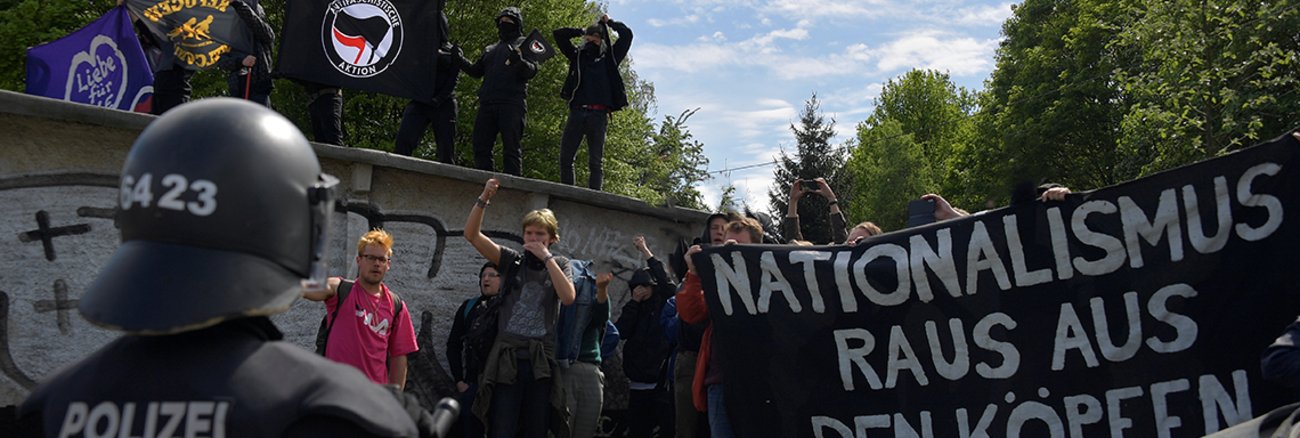 Antifa-Demonstranten halten Fahnen hoch, ein Polizist mit Helm steht ihnen gegenüber 