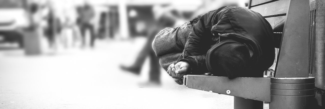 Eine wohnungslose Person liegt auf einer Bank der Straßenmöblierung einer Innenstadt und schläft. Foto: © istock.com/Srdjanns74
