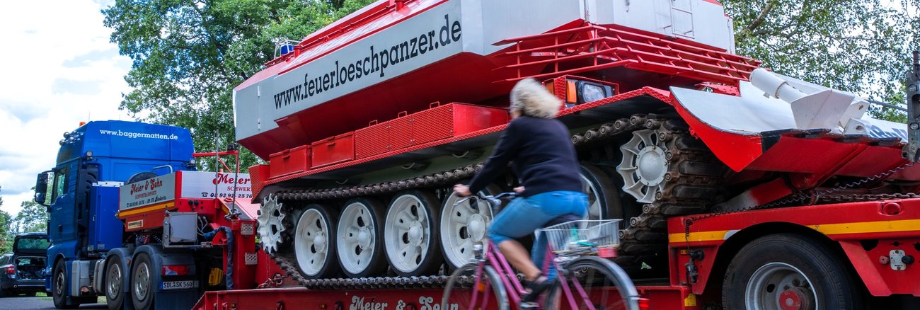 Waldbrand in Lübtheen in Mecklenburg-Vorpommern: Eine Frau fährt mit einem Fahrrad an einem Tieflader mit einem Feuerlöschpanzer vorbei © dpa/Jens Büttner