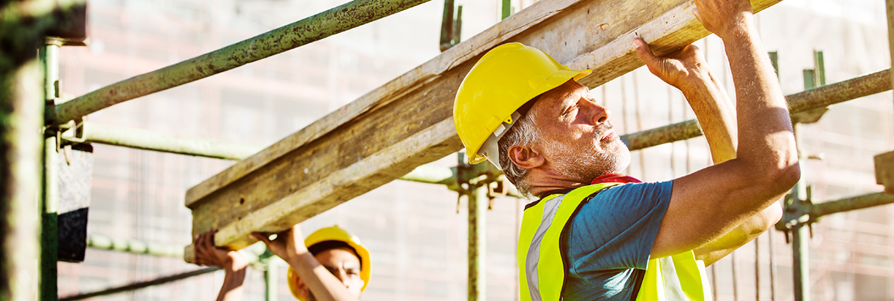 Zwei Arbeiter mit Helm und Schutzweste stemmen einen Stahlträger auf einer Baustelle. Foto: © istock.com/Morsa Images