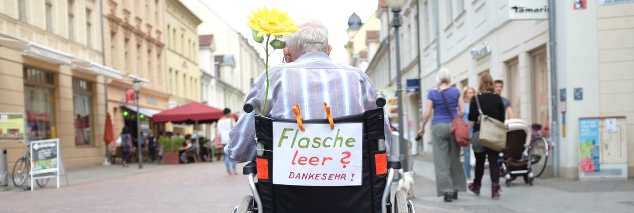 Ein alter Mann im Rollstuhl mit einem Zettel »Flasche leer? Danke sehr!« sammelt in einer Fußgängerzone leere Flaschen © dpa/Kay Nietfeld