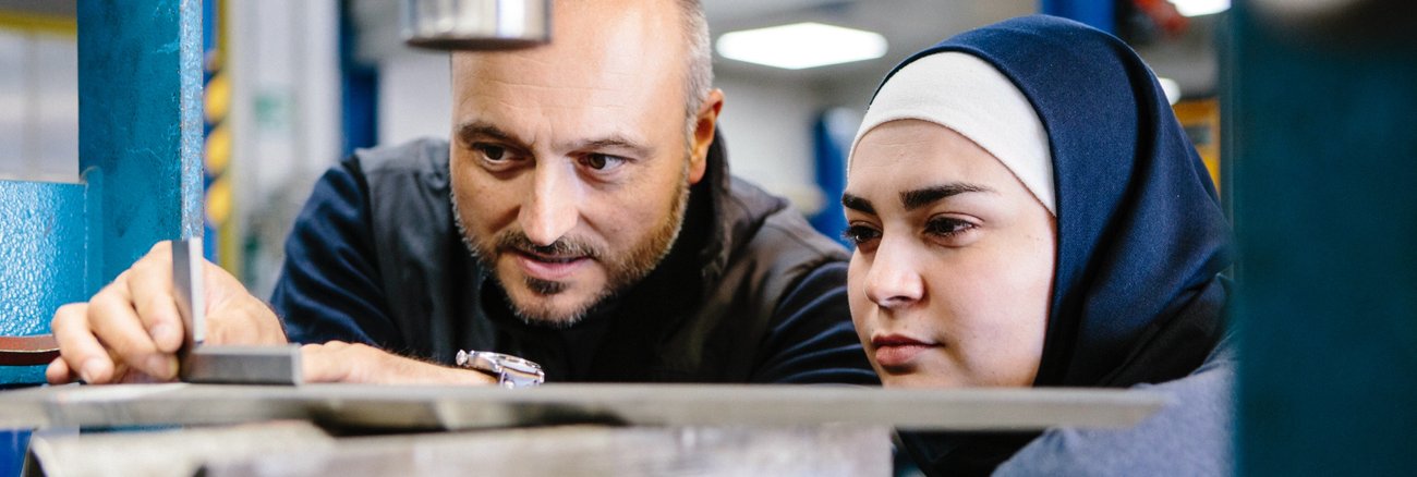 Eine Muslima mit Kopftuch und in Latzhose arbeitet zusammen mit einem Kollegen mit Messschieber an einer Werkzeugmaschine © iStock/fotografixx