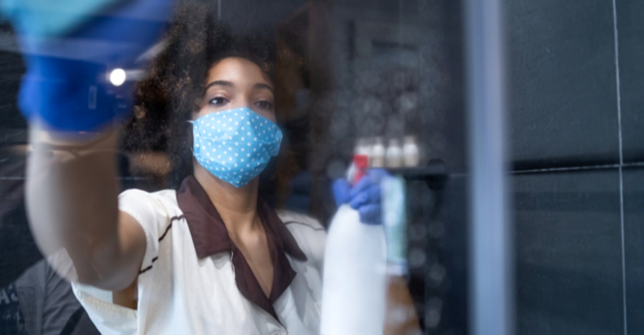 Eine Putzfrau mit Maske, Gummihandschuhen und einer Sprühflasche in der Hand reinigt einen Spiegel © iStock/valentinrussanov