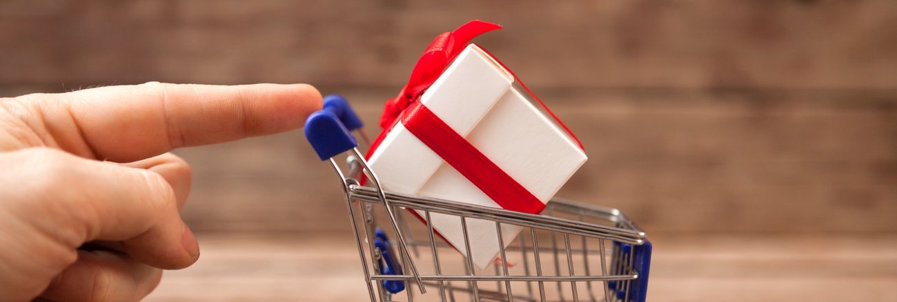 Ein Finger berührt einen Miniatur-Einkaufswagen mit einem Mini-Paket und roter Schleifer © iStock/123ducu