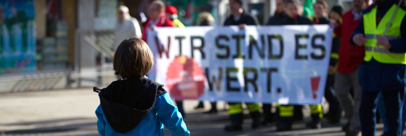 Ein kleiner Junge mit Laufrad steht vor Demonstranten, die eine Banner halten mit der Aufschrift: Wir sind es wert.  © picture alliance/dpa|Uwe Zucchi