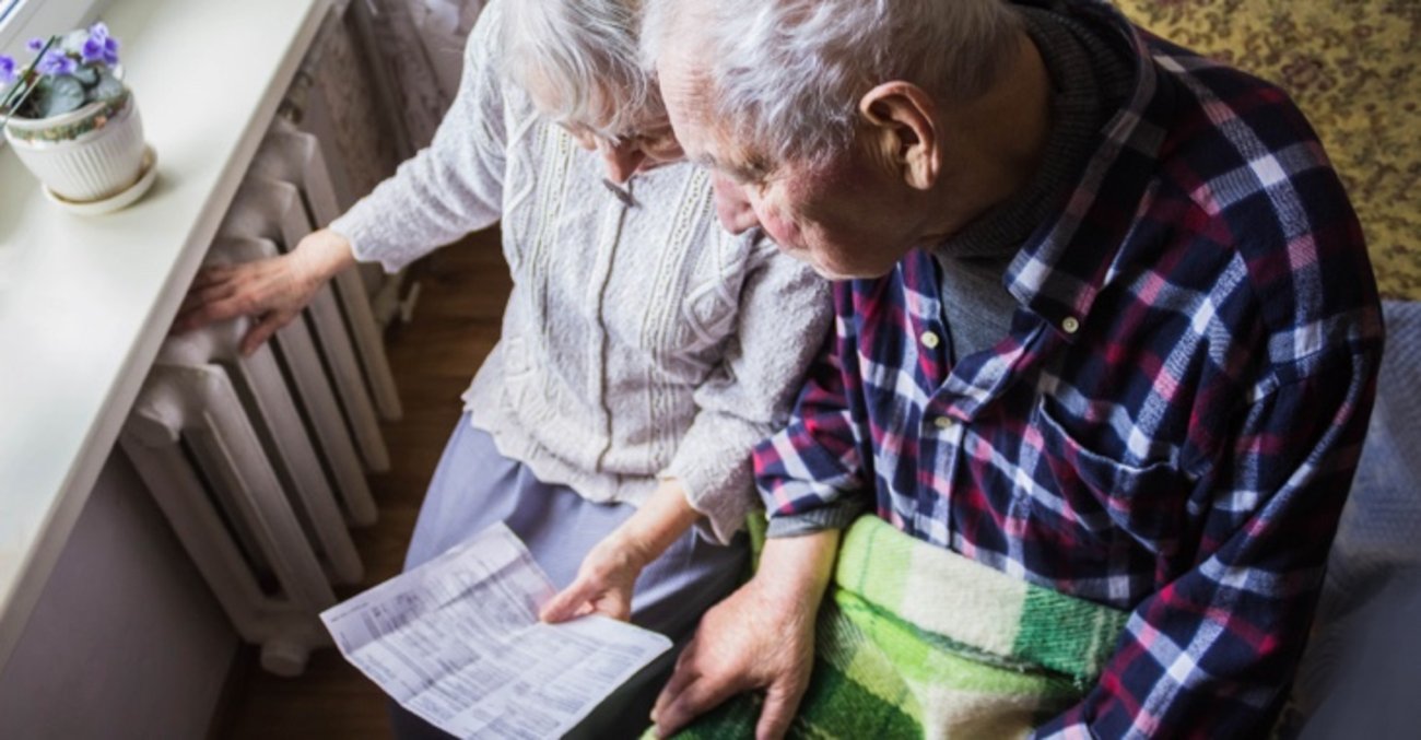 Eine ältere Frau hält eine Rechnung und fasst auf einen Heizkörper, daneben ein älterer Mann mit einer Decke auf den Beinen © iStock/Marina113