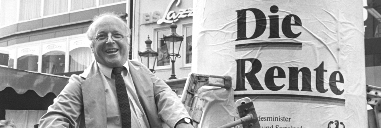Norbert Blüm am 21. April 1986 mit einem Besen an einer Litfaßsäule mit dem Plakat »Denn eins ist sicher: Die Rente« © picture alliance/Peter Popp