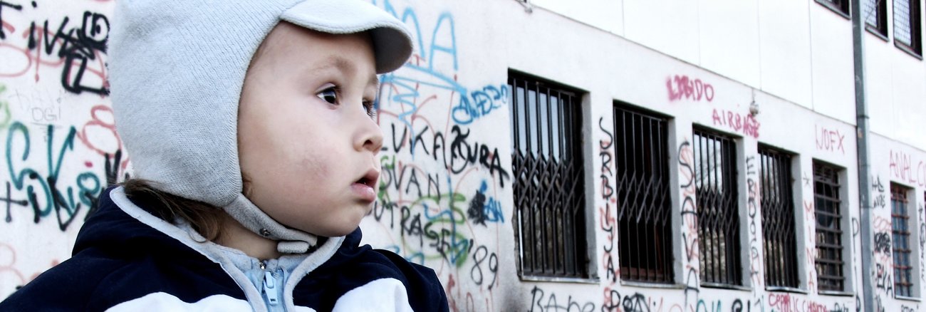 Ein Kind vor einem Haus mit Graffiti an der Wand und Gittern vor den Fenstern © iStock/brunette