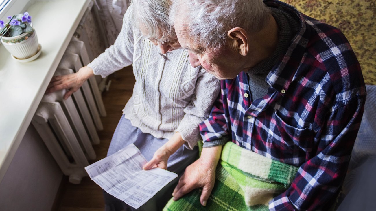 Eine ältere Frau hält eine Rechnung und fasst auf einen Heizkörper, daneben ein älterer Mann mit einer Decke auf den Beinen © iStock/Marina113