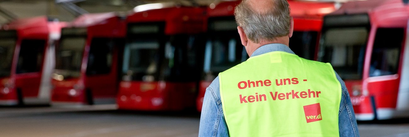 Ein Mann vor roten Straßenbahnen trägt eine gelbe Warnweste mit der Aufschrift: Ohne uns kein Verkehr © picture alliance/dpa|Daniel Karmann