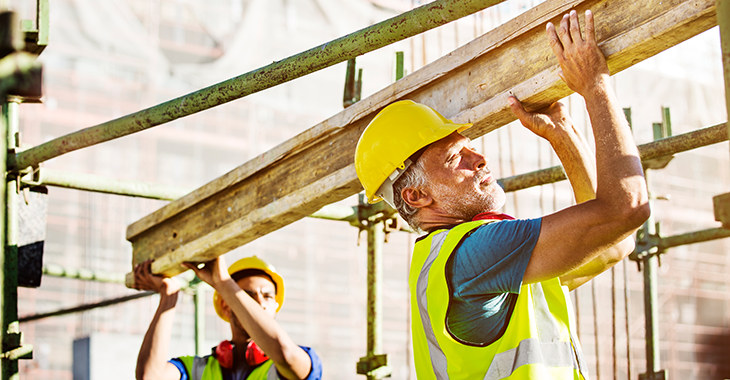 Zwei Arbeiter mit Helm und Schutzweste stemmen einen Träger auf einer Baustelle. Foto: © istock.com/Morsa Images