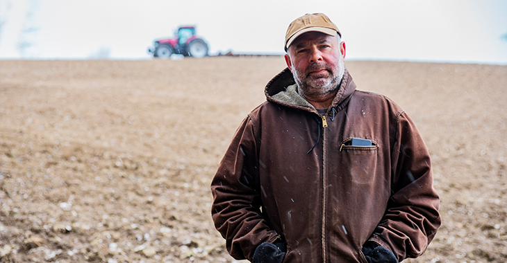 Ein Landwirt steht auf einem winterlichen Acker, über den unscharf im Hintergrund ein Traktor mit Arbeitsgerät fährt. Foto: © istock.com/Fertnig