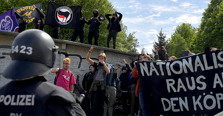 Antifa und Polizei stehen sich gegenüber @Reuters/Matthias Rietschel Antifa-Demonstranten halten Fahnen hoch, ein Polizist mit Helm steht ihnen gegenüber