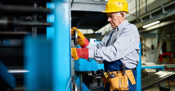 Ein älterer Arbeiter mit gelbem Schutzhelm schraubt an einer blauen Maschinen © iStock/SeventyFour