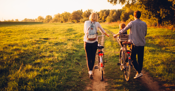 Junge Eltern mit Kind schieben zwei Fahrräder über ein Feld © iStock/AleksandarNakic Junge Eltern mit Kind schieben zwei Fahrräder über ein Feld © iStock/AleksandarNakic