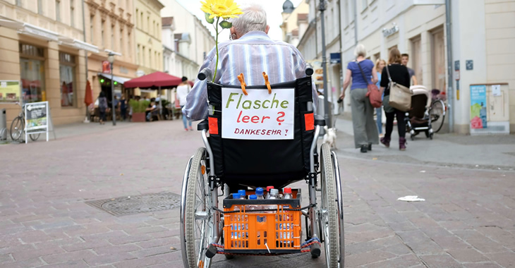 Ein älterer Mann in einer Fußgängerzone hat an seinem Rollstuhl einen Zettel befestigt mit der Aufschrift »Flasche leer? Danke sehr!« befestigt © dpa
