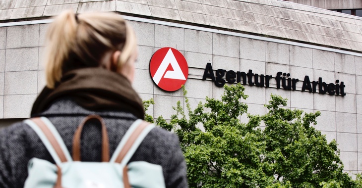 Frau mit Rucksack vor einem Gebäude mit dem Logo und der Aufschrift Agentur für Arbeit © picture alliance/imageBROKER|Jan Tepass