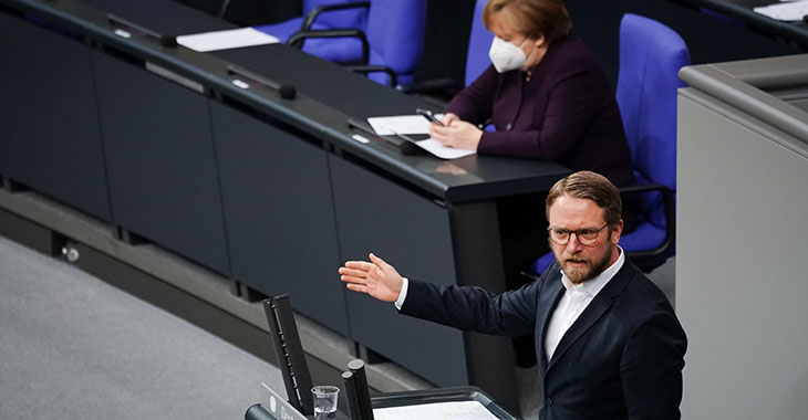 Jan Korte spricht im Plenum mit Angela Merkel im Hintergrund