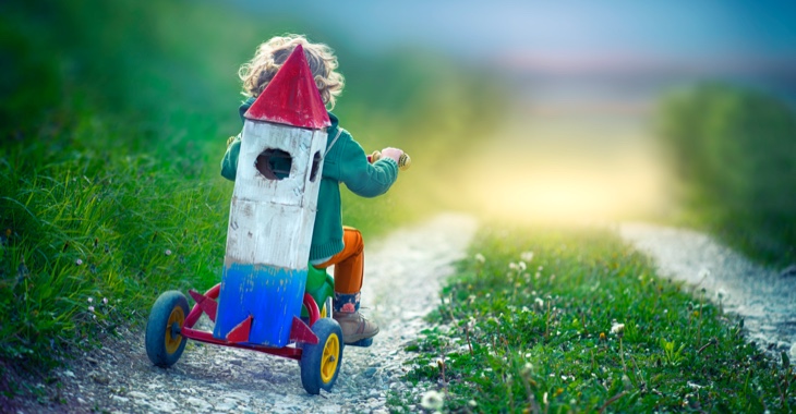 Ein Kind fährt mit einem Dreirad auf einen Feldweg mit einer aus Pappe gebastelten Weltraum-Rakete auf den Rück geschnallt © iStock/oops7